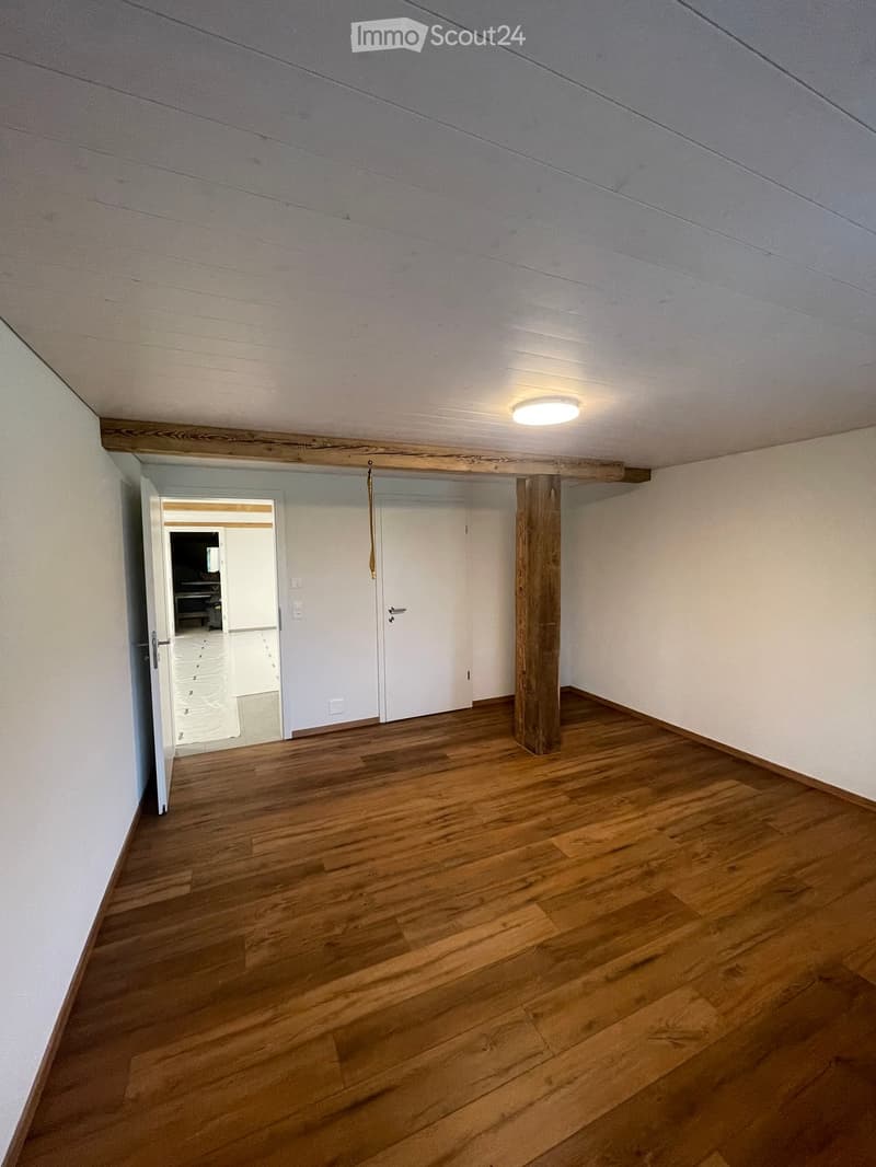 Empty room with wooden flooring, white walls, and a single ceiling light. A wooden post stands in the center, and an open doorway leads to another room.