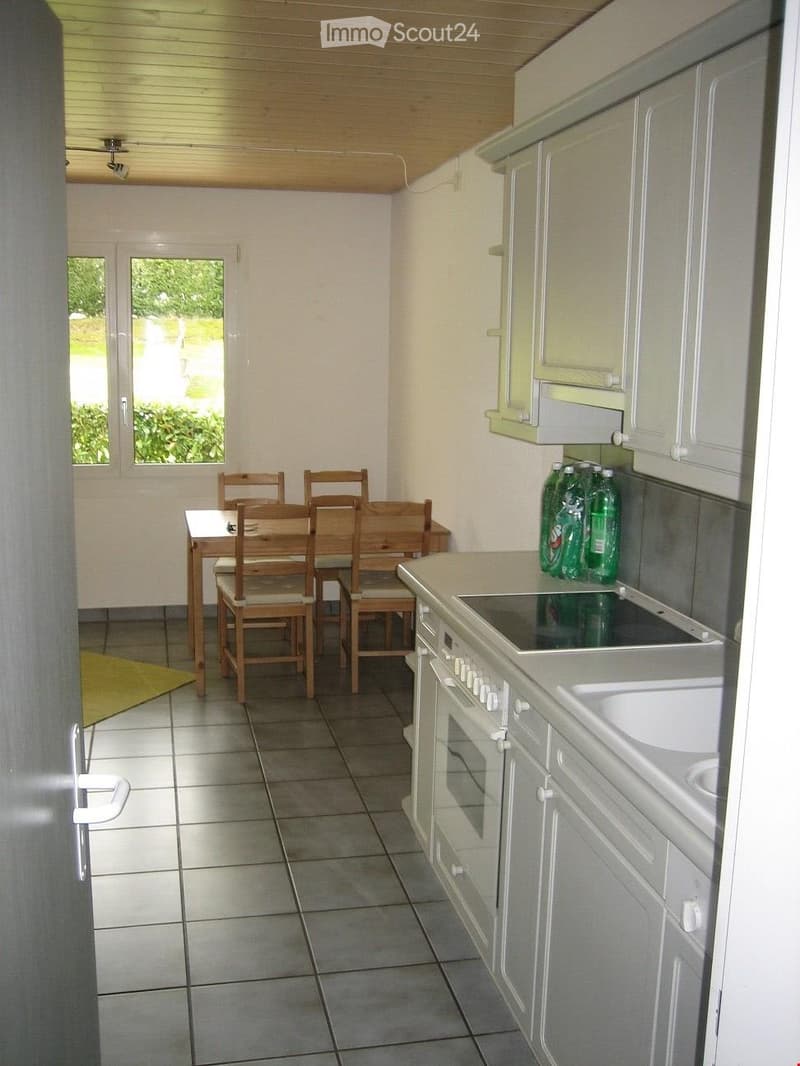 A modern kitchen with white cabinets, a tiled floor, and a dining area with a wooden table and chairs. There is an oven, sink, and several green bottles on the counter.