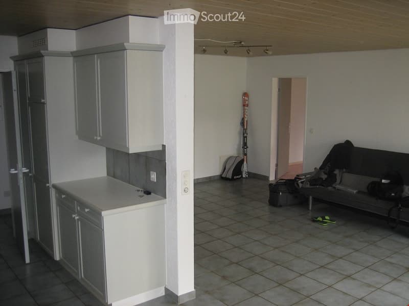 A small, open-plan space with a kitchen area on the left and a living area on the right. The kitchen features white cabinets and a countertop, while the living area includes a sofa, a bench, skis, a backpack, and luggage.