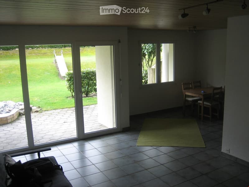 Bright living room with large sliding glass doors opening to a garden, tiled floor, green rug, sofa, dining table with chairs.