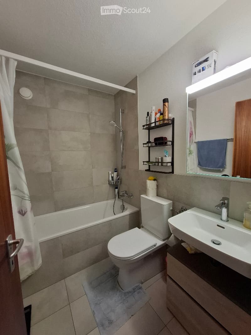 Modern bathroom with a bathtub, toilet, sink, and a mirror. A black shelf holds various toiletries, and a blue towel hangs on a rack. The shower area features a glass enclosure and a showerhead.