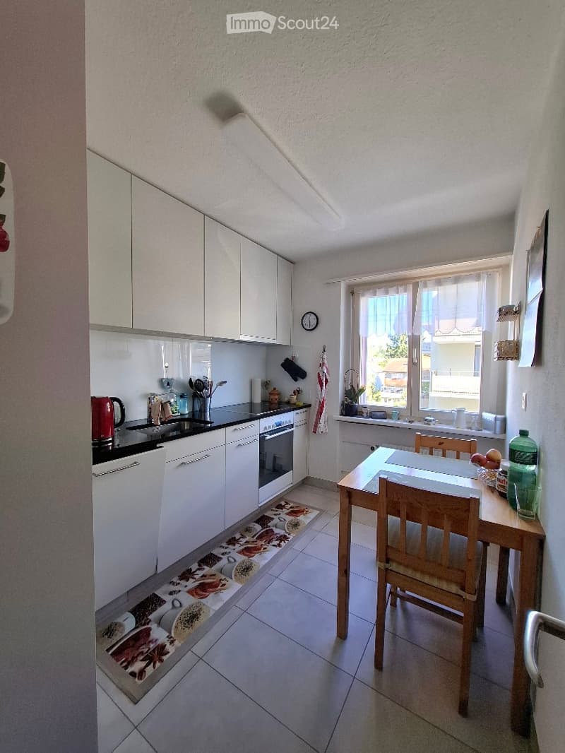 Modern kitchen with white cabinets, black countertops, and a small dining area with a wooden table and chairs. The kitchen features a sink, oven, and various kitchen utensils.