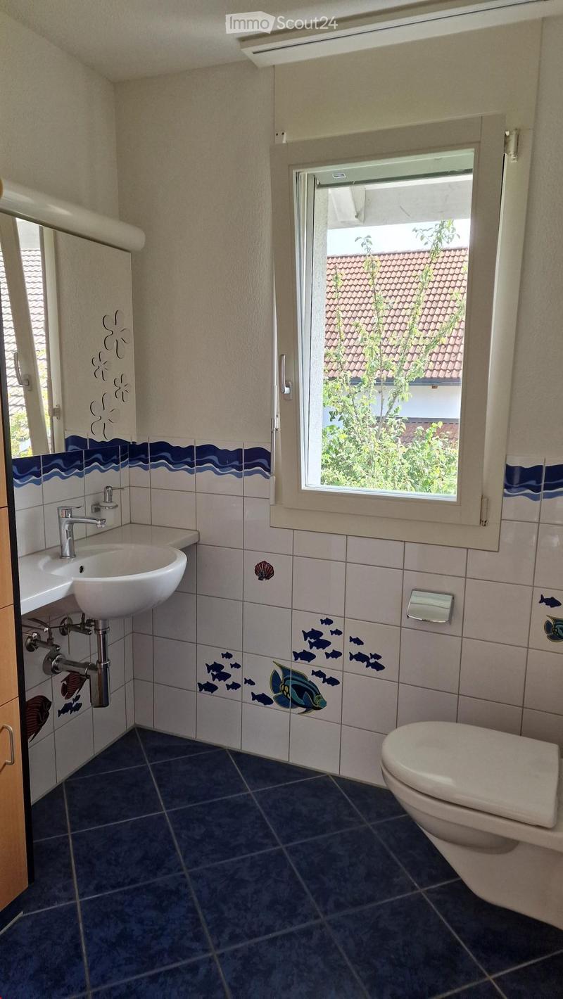 Bathroom with white tiles and blue tile flooring, a toilet, a white sink with faucet, a mirror with fish designs, and a window with a view of trees and a house.