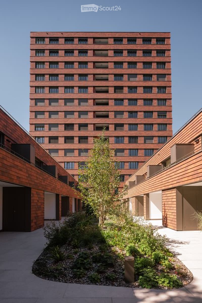Modern multi-story building with a central courtyard featuring a small tree and shrubs. The building has a uniform brick facade with numerous windows and a flat roof.