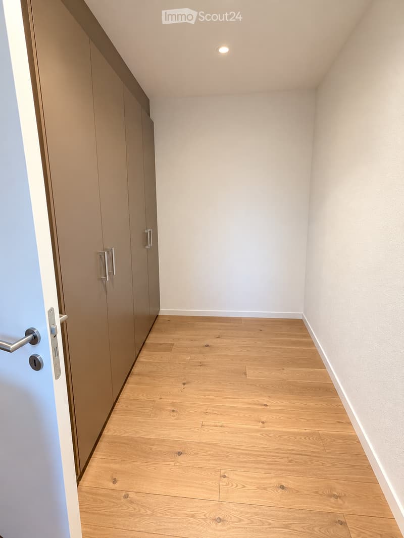 Empty room with light wooden flooring, white walls, and built-in closet with silver handles. The room has a recessed ceiling light and a door visible on the left side.