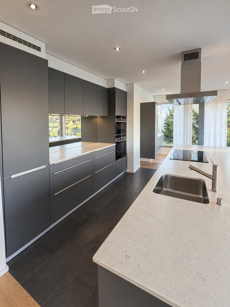 Modern kitchen with dark grey cabinets, white marble countertop, stainless steel sink and faucet, built-in oven, and large windows offering a view of greenery.