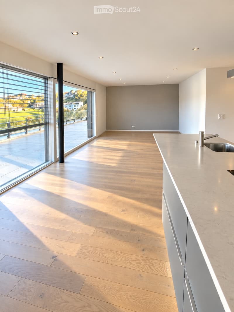 Modern, minimalist kitchen with light wooden flooring, white cabinetry, and a sleek countertop. Large windows offer a scenic view of the outdoors.