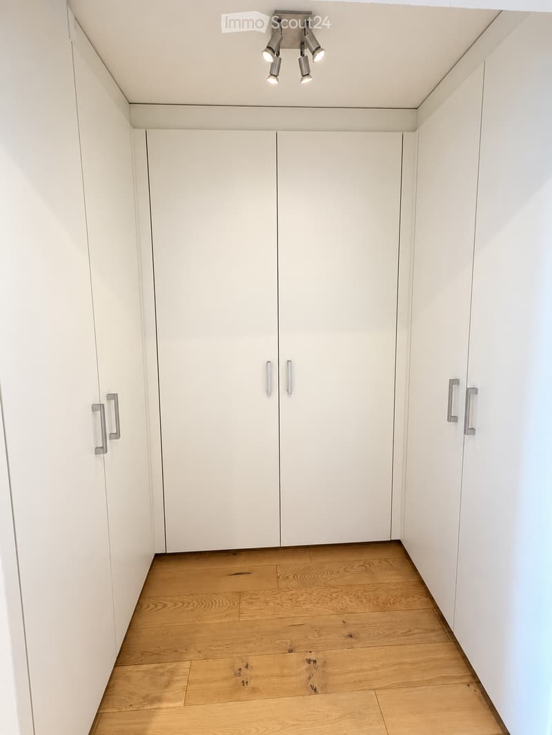 Empty room with white walls, light wooden flooring, and built-in white cabinets with silver handles. A recessed ceiling light fixture is visible.