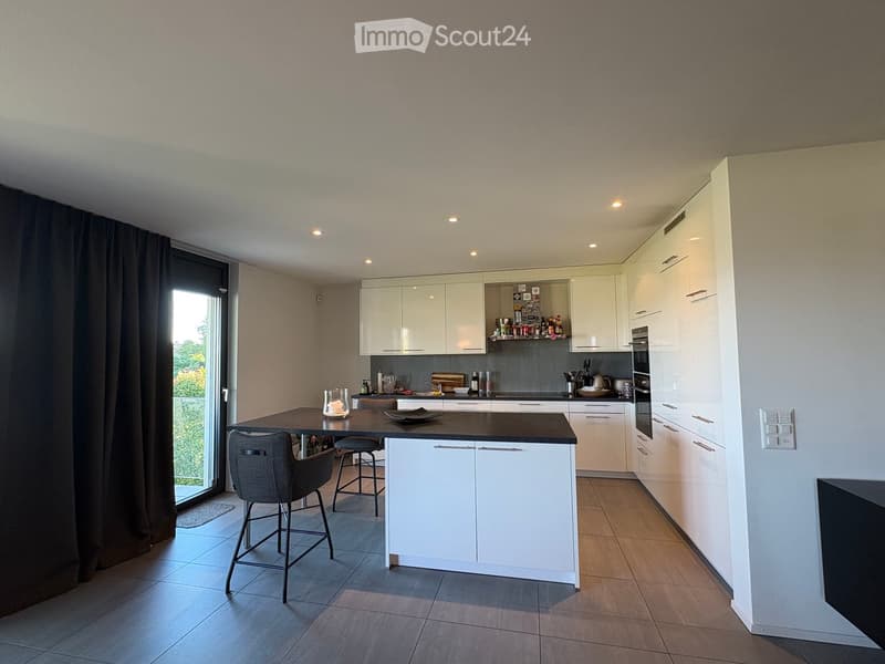 Modern kitchen with white cabinets, a central island, and bar stools. Features an oven, countertop items, and a large window with dark curtains.