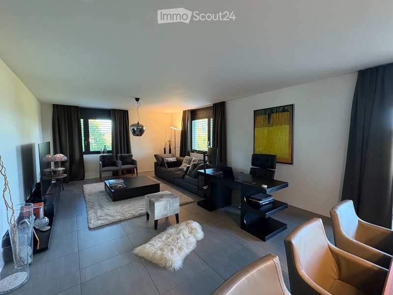 Modern living room with a sofa, coffee table, chairs, a small desk with a laptop, and decorative items including a vase and lamps. The space features large windows with curtains and a cozy rug on the floor.
