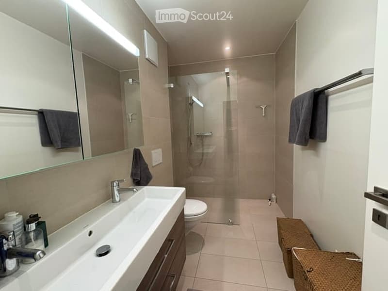 Modern bathroom with a large mirror, sink, toilet, and glass-enclosed shower. Towels are neatly hung on racks, and a wicker basket is placed on the floor.