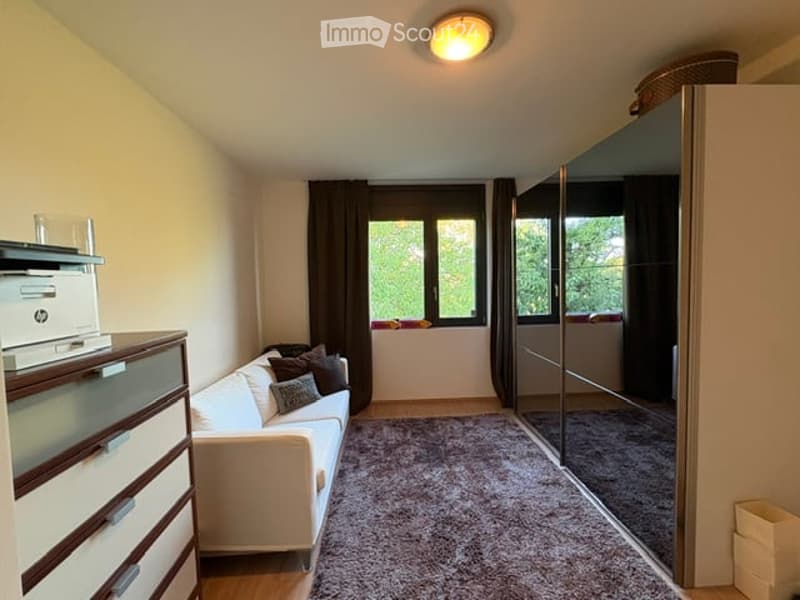 A small, well-lit room with a white sofa, a wooden dresser, a large mirror, and a printer on a shelf. The room features a carpeted floor and large windows with dark curtains.