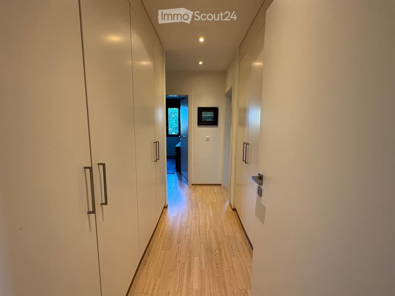 A modern hallway with light wooden flooring, white walls, built-in wardrobes with silver handles, recessed ceiling lights, and a small framed picture on the wall.