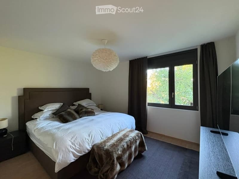 Modern bedroom with a large bed, nightstand, lamp, chair, and a small table near the window with curtains.