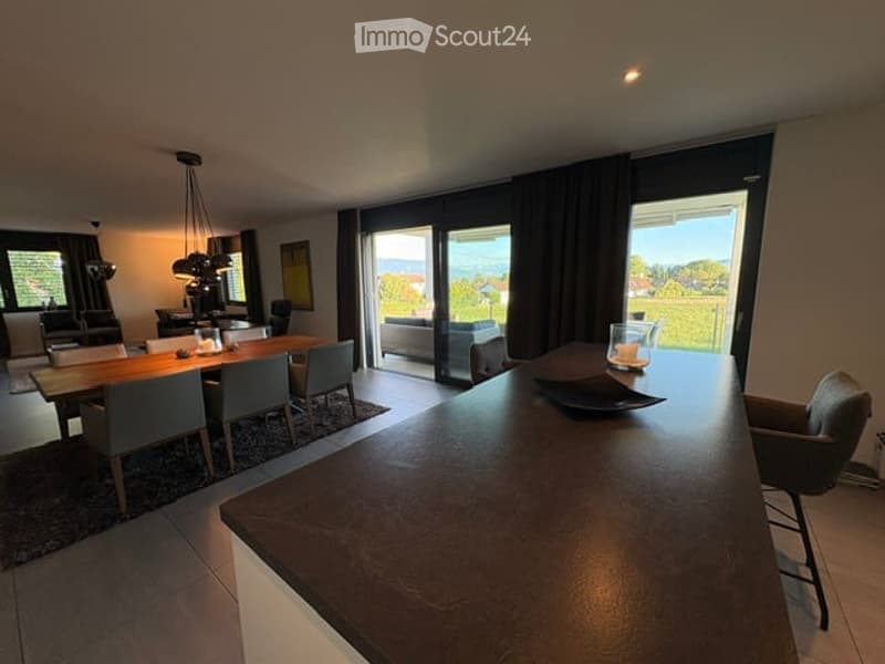 Modern open-plan living and dining area with large windows offering a scenic view, featuring a wooden dining table, chairs, a sofa, a coffee table, and a bar area with a stool.