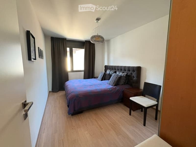 Cozy bedroom with wooden flooring, a large bed with a dark headboard and colorful bedding, a nightstand, a chair, and dark curtains on the window. A decorative chandelier hangs from the ceiling.