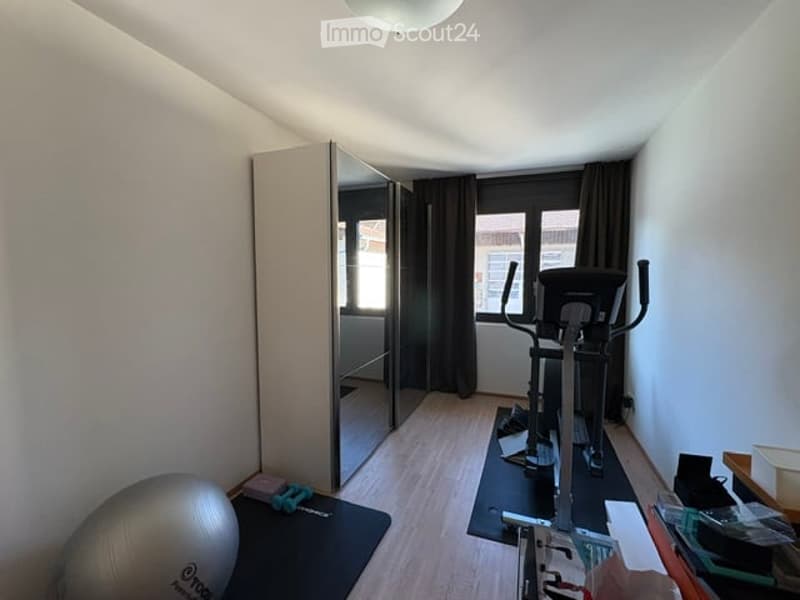 A well-lit room with wooden flooring, featuring an exercise bike, a large mirror, a wardrobe, and a fitness ball on a mat.