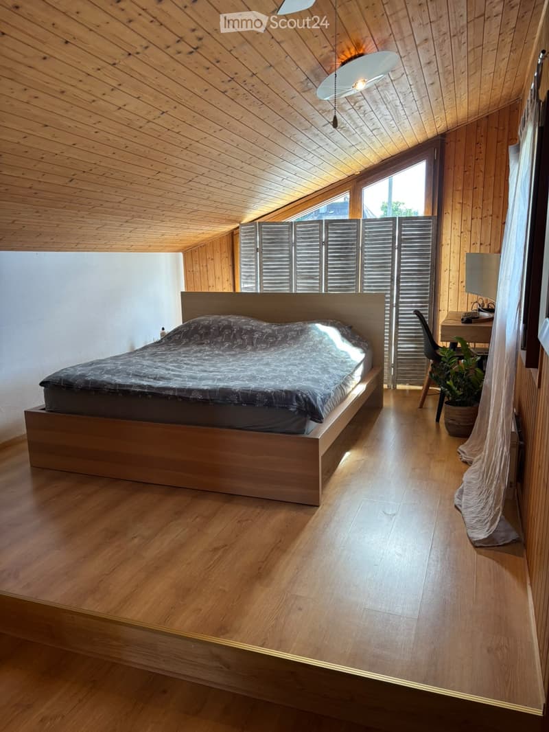 Cozy attic bedroom with wooden floors and slanted wooden ceiling, featuring a large bed, a small desk with a chair, and a potted plant near the window.