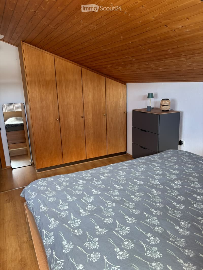 Cozy bedroom with wooden floor, slanted wooden ceiling, large wardrobe, bed with grey floral duvet, nightstand with lamp and decorative item.
