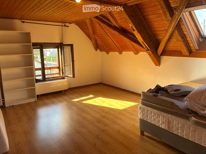 Bright bedroom with wooden floors, slanted wooden ceiling, large window with open shutters, bed with pillows, and a bookshelf.