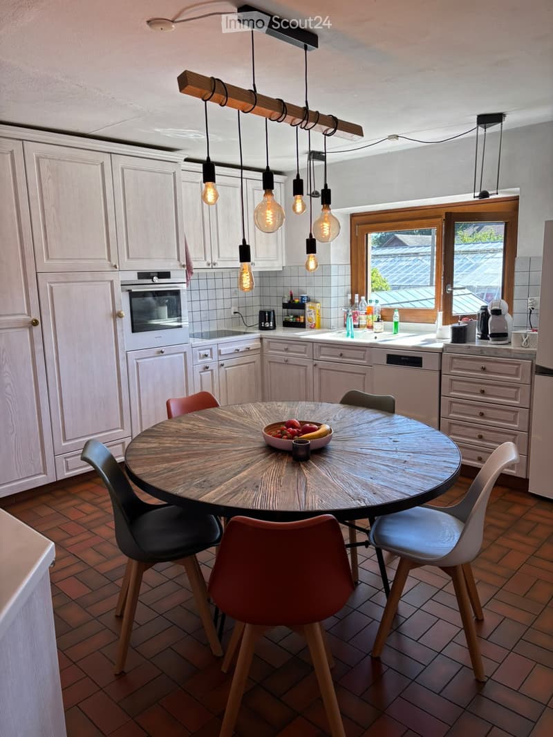 Modern kitchen with light-colored cabinets, a round wooden dining table with four chairs, and various appliances including an oven, dishwasher, and refrigerator. The room features pendant lights and a window with a view of a roof.