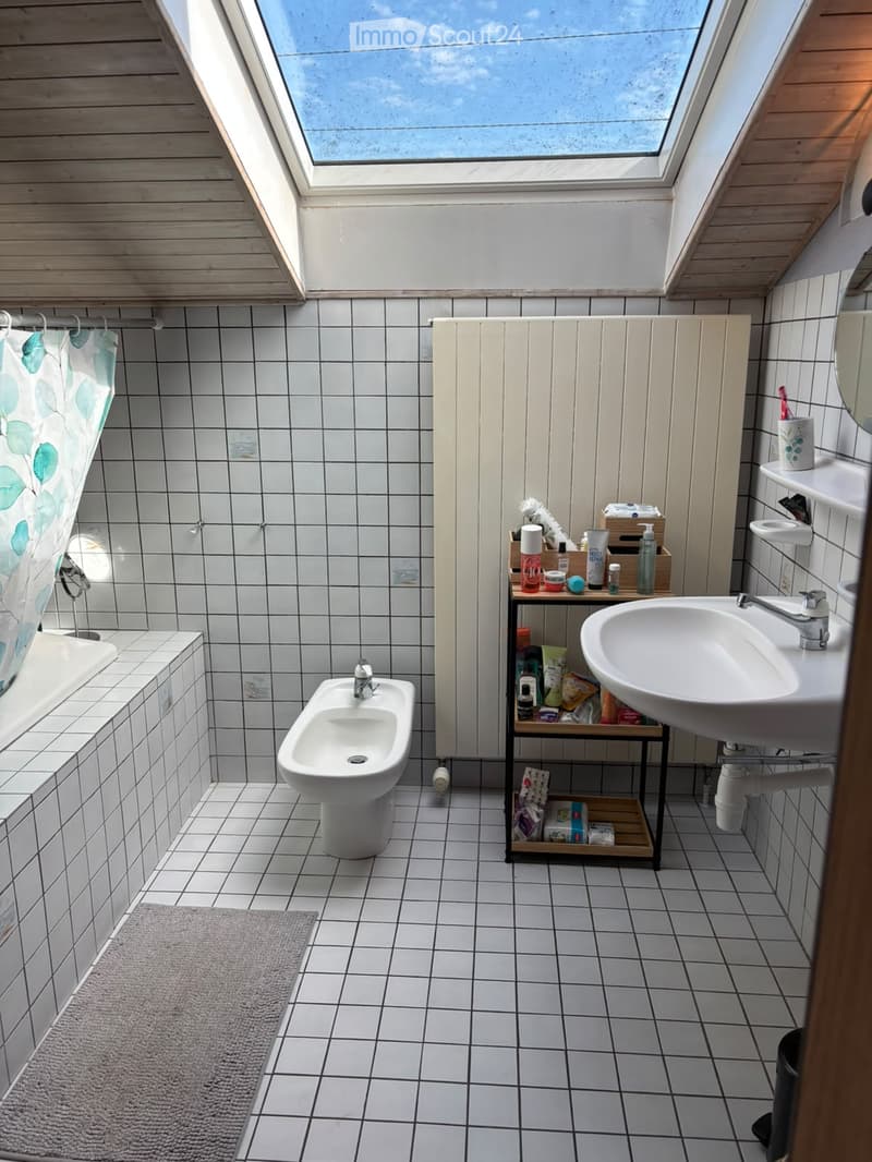 Bright, modern bathroom with white tiles, a bathtub, bidet, sink, and a small shelf holding toiletries. The room features a skylight and a grey bath mat.