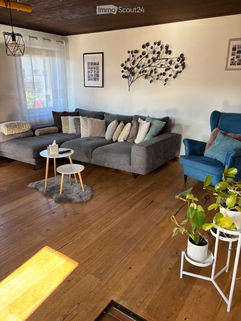 Modern living room with wooden flooring, grey sectional sofa, blue armchair, small coffee tables, potted plants, and decorative wall art.