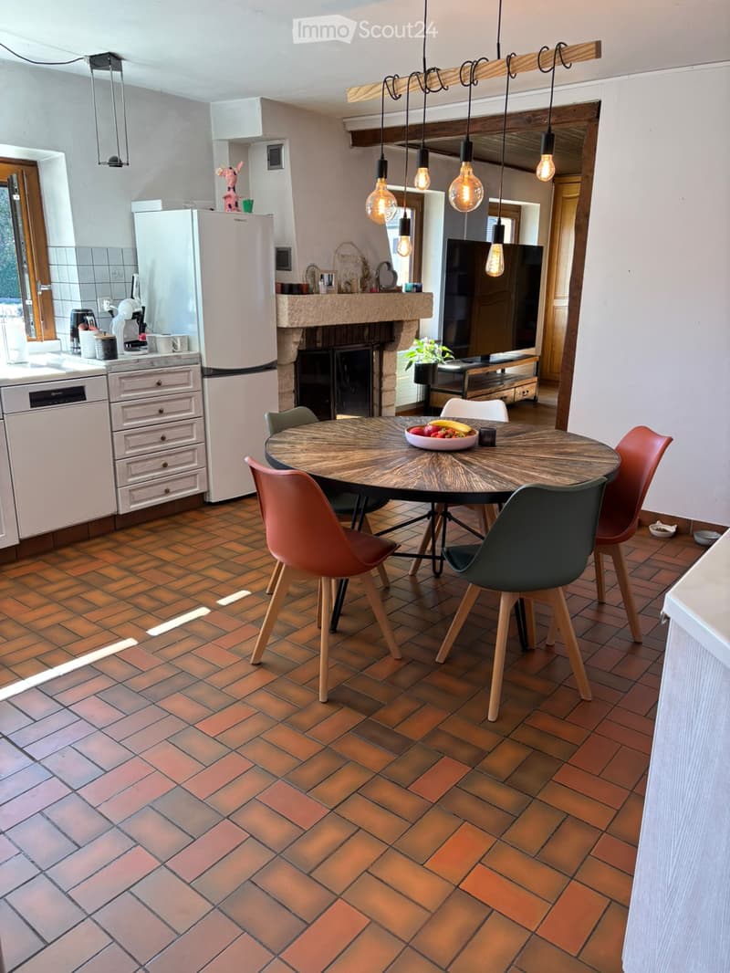 A modern kitchen with a dining area, featuring a round wooden dining table with colorful chairs, a refrigerator, dishwasher, and a fireplace. The space is illuminated by pendant lights and has a tiled floor.