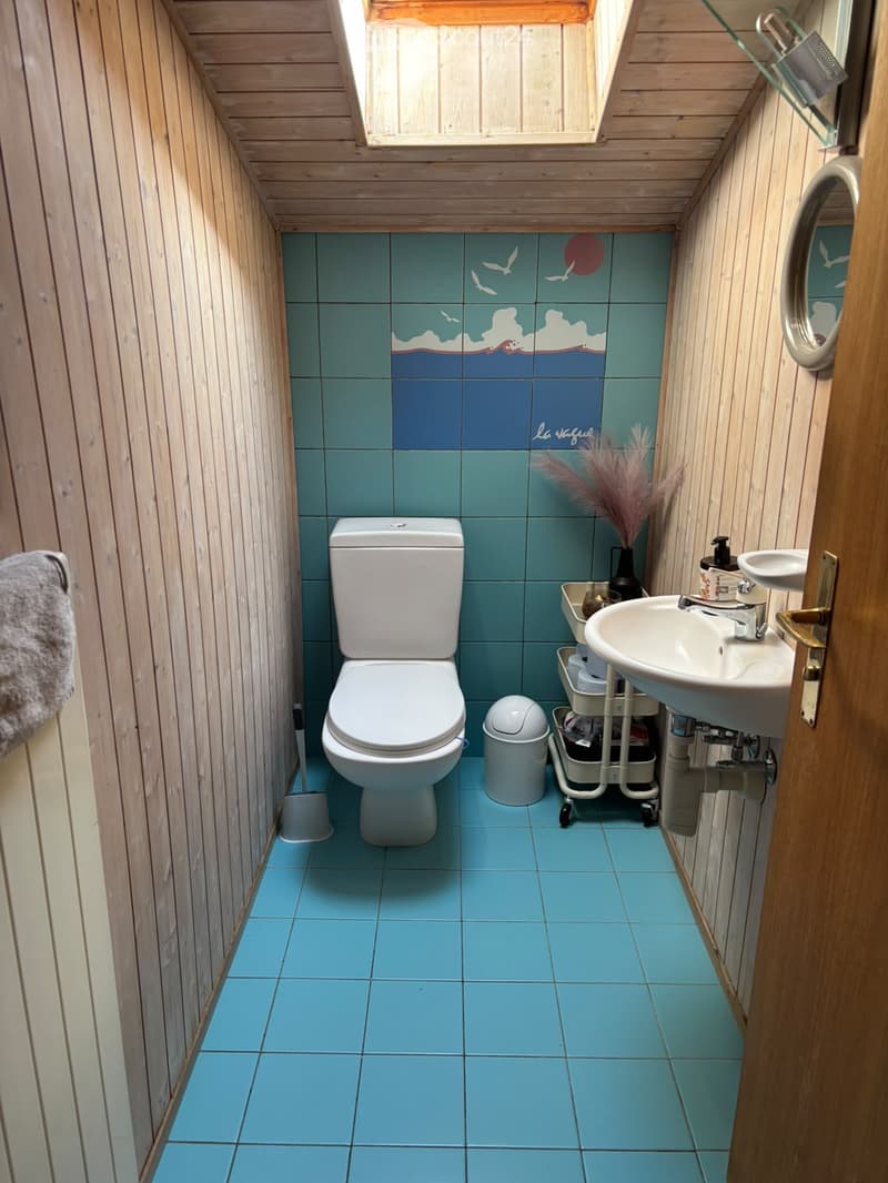 Cozy bathroom with light wood paneling, blue tiles, a toilet, sink, mirror, and decorative vase.