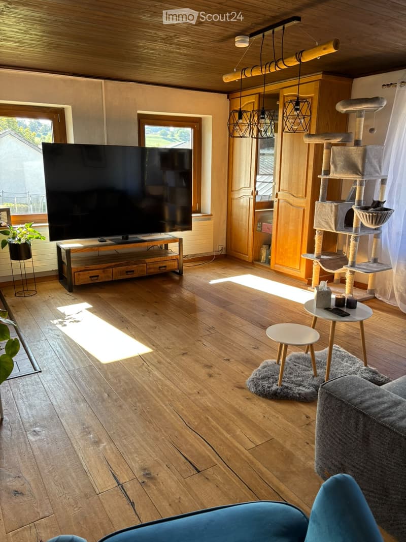Living room with wooden flooring, large windows, a grey sofa, a cat tree, a television on a wooden stand, and a small round table with a remote control and candles.