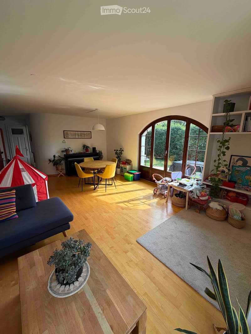 Bright living room with wooden flooring, large arched windows, sofa, coffee table, dining table with chairs, piano, and various plants and toys.