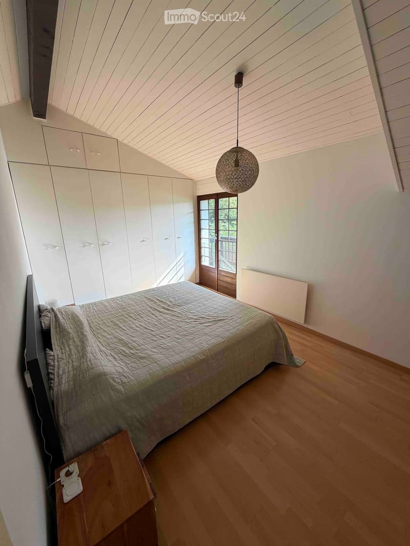 Modern bedroom with wooden flooring, a large bed with grey bedding, built-in white wardrobes, a pendant light fixture, and a nightstand with a lamp.