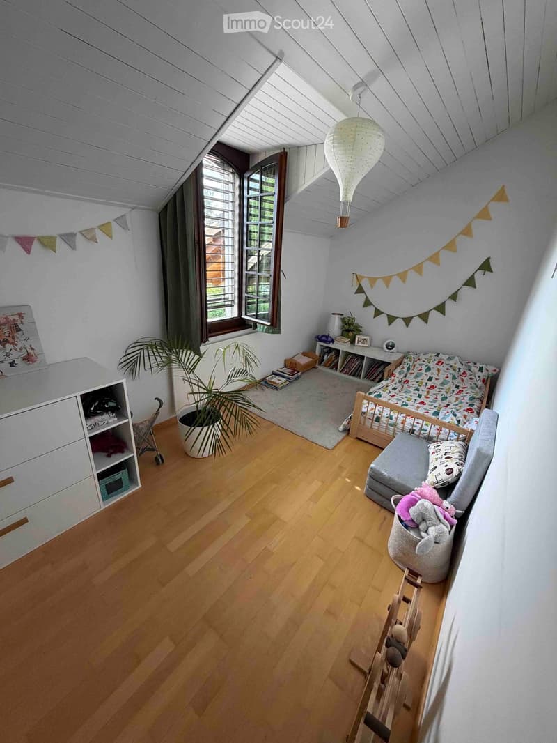 Cozy attic bedroom with wooden floors, slanted ceilings, large windows, a small bed, a sofa, a shelf, a plant, and decorative items.