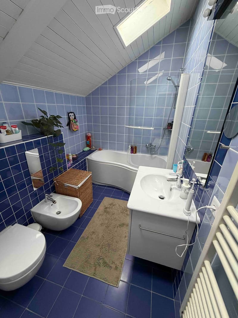 Modern bathroom with blue tiles, white fixtures, a bathtub, sink, toilet, and a small shower area. Includes a wicker basket, plant, and a bathroom rug.