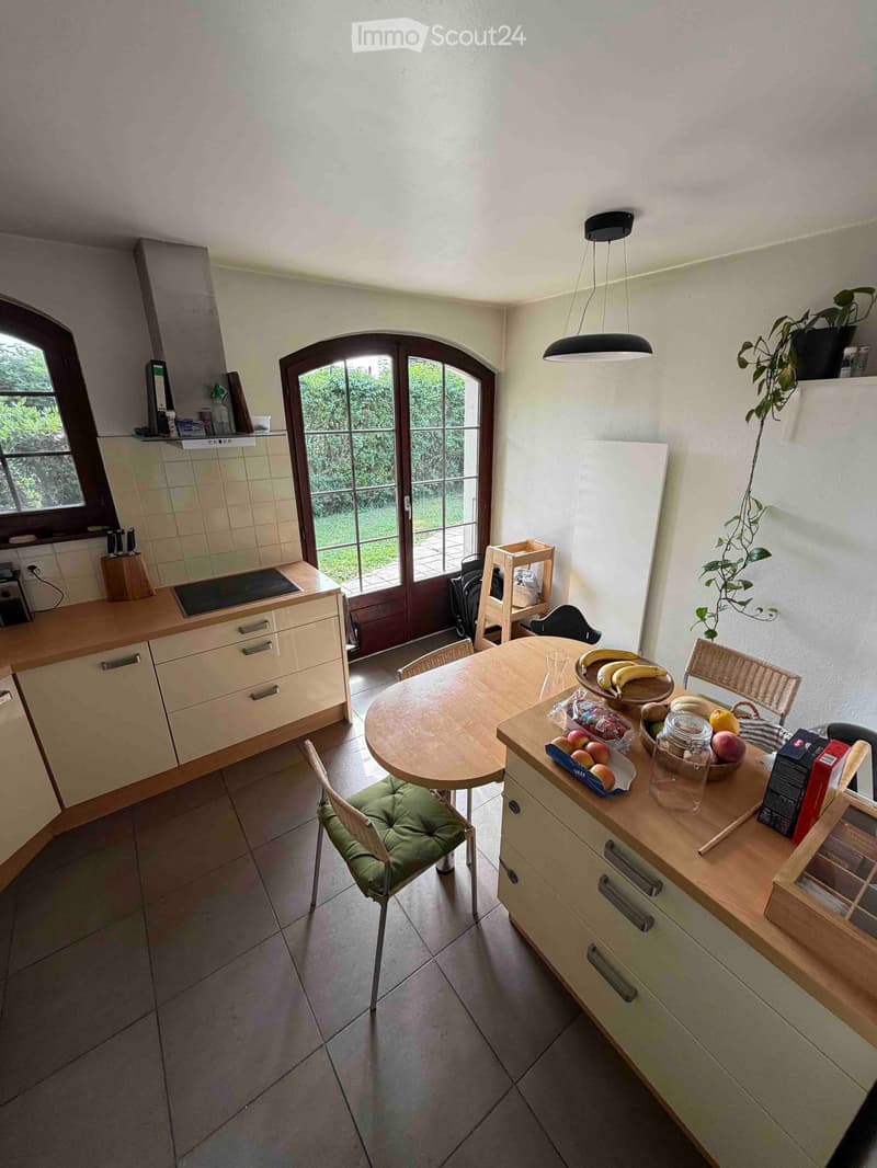 Bright kitchen with light-colored cabinets, an induction stove, a round wooden dining table with chairs, and a countertop with fruit and a potted plant. Large arched windows provide natural light.