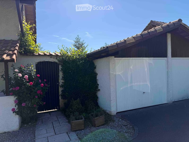 A two-story building with a tiled roof, white walls, and a gated entrance. The entrance features a dark wooden door surrounded by climbing plants and flowers. A white garage door is visible to the right, and small wooden planters are placed near the entra