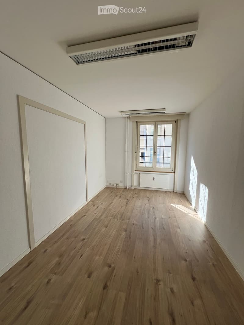 Empty room with wooden flooring, white walls, and a large window with multiple panes. The room has a door on the left and a radiator below the window.