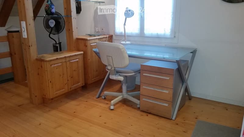 A modern, well-lit room with wooden flooring, a glass desk, a white ergonomic chair, a fan, and a wooden cabinet.