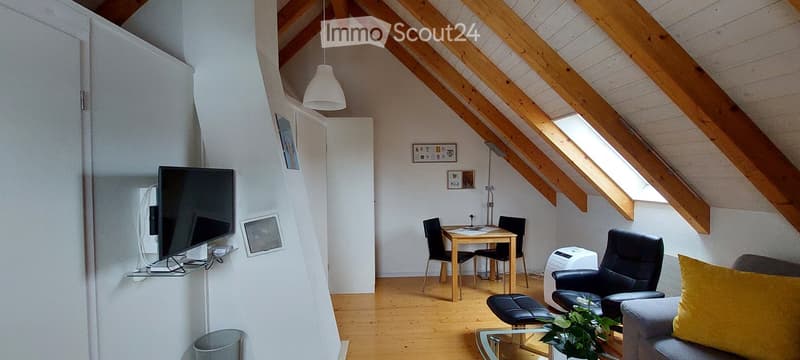 Bright attic room with wooden beams, white walls, and wooden flooring. Features a sofa, armchair, coffee table, dining table with chairs, and a TV on a stand.