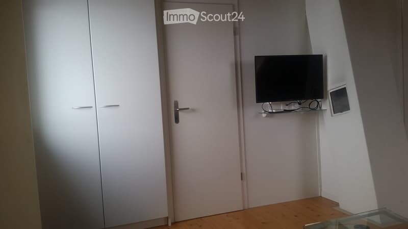 A small, modern room with a TV mounted on the wall, a glass table, and a white cabinet with a door.