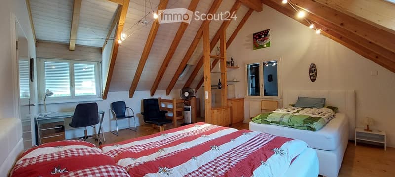 Cozy attic bedroom with wooden beams, two beds, desks, chairs, and a fan. Large windows provide natural light.