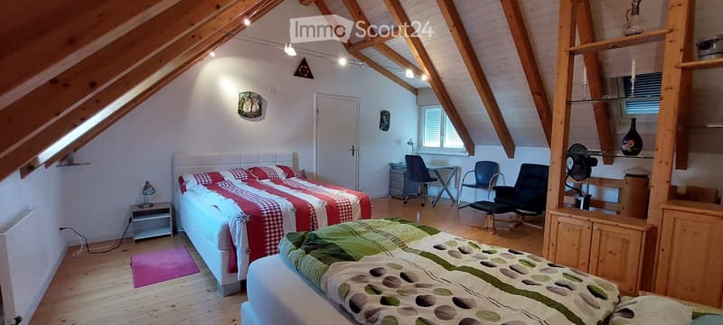 Cozy attic bedroom with wooden beams, two beds, a desk area with chairs, a nightstand lamp, and built-in shelving with decorative items.