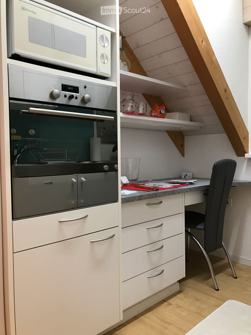 A compact, modern kitchen with white cabinets, stainless steel appliances, and a small desk area with a chair.