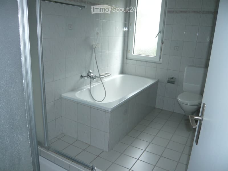 Modern bathroom with a bathtub, toilet, and a glass-enclosed shower area. The room features white tiled walls and floor, with a window providing natural light.