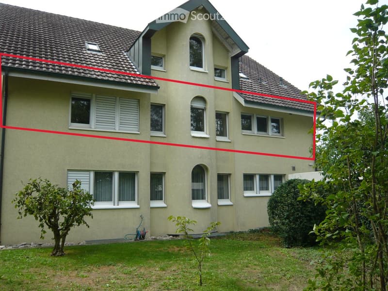 Two-story house with beige siding, multiple windows, and a tiled roof. The house features a small garden with grass, trees, and shrubs in the foreground.