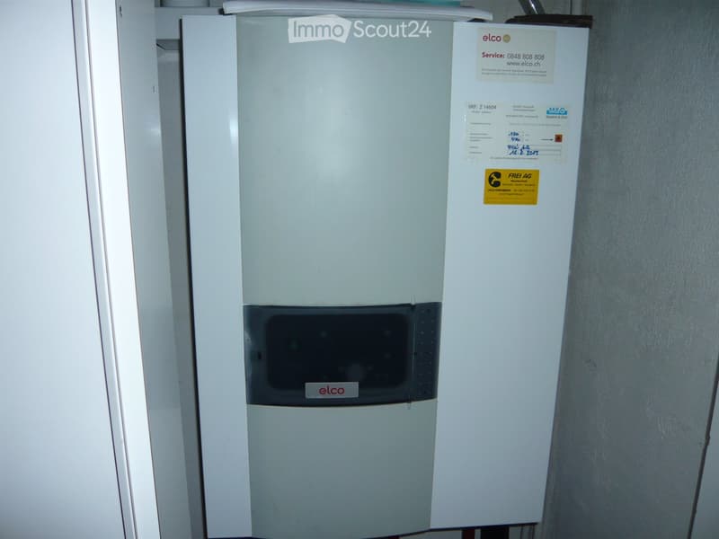 A white refrigerator with a control panel and various labels and stickers on the front, placed in a room with a white wall and a white door frame visible in the background.