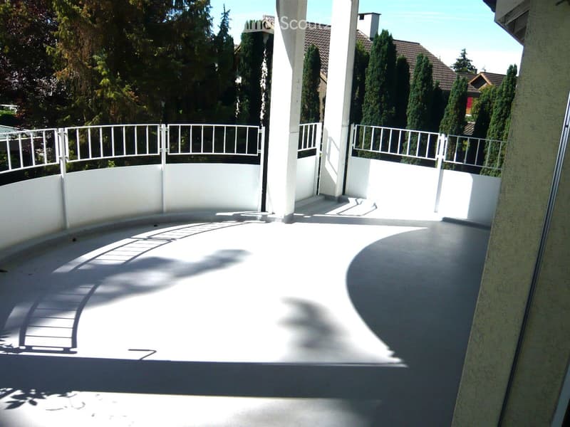 A modern balcony with white railings, a smooth concrete floor, and a view of surrounding trees and houses.