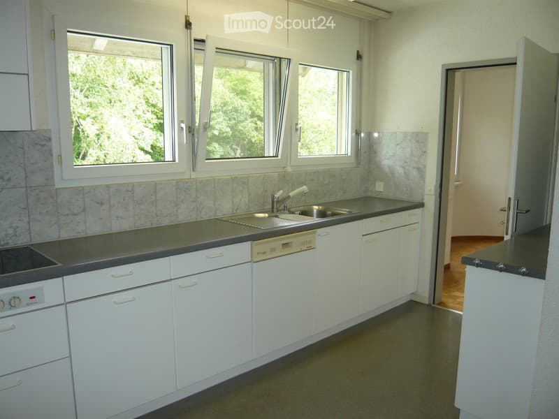 Modern kitchen with white cabinets, stainless steel sink, dishwasher, and oven. The countertops are dark grey, and the backsplash features a marble tile design. Three windows with white frames allow natural light to enter.