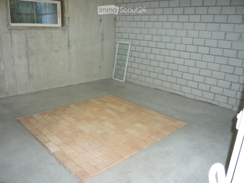 Empty room with concrete floor, exposed brick wall, and a single window. A small rug is placed on the floor, and a white frame leans against the wall.