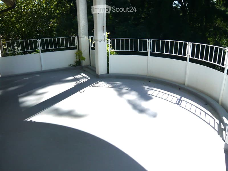 Empty balcony with white railings, concrete floor, and lush green foliage in the background.